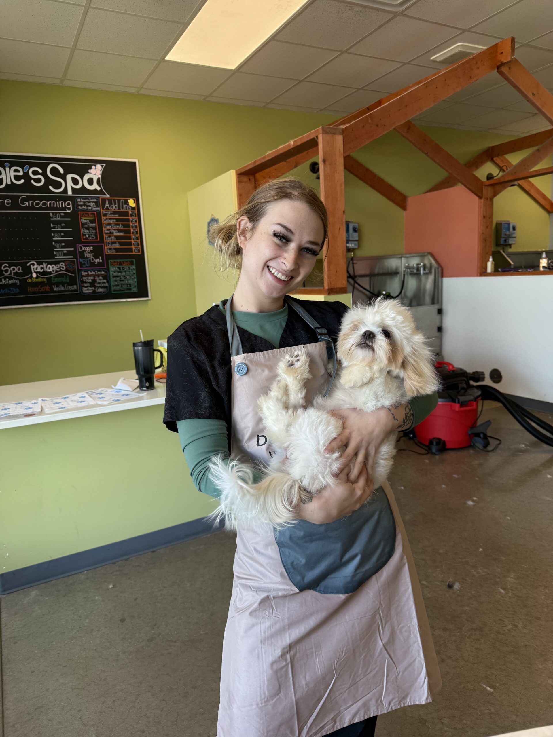 Smiling pet groomer in a pink apron holding a fluffy white dog inside a grooming salon with a chalkboard menu in the background.
